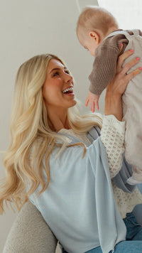 A woman sitting in a modern white armchair holds a baby wrapped in a light gray blanket. The room has a large oval window, a full-length mirror, and a concrete floor.