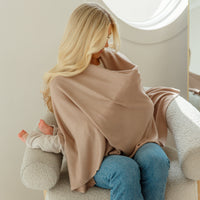 A woman sits on a cream boucle armchair, seen from behind, wearing a large taupe knit baby blanket or shawl over her shoulders and blue jeans. A baby is nestled in her lap.